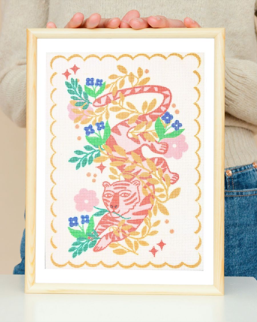 A needlepoint kit depicting a tiger with flowers and plants in vibrant colors, held in a frame.