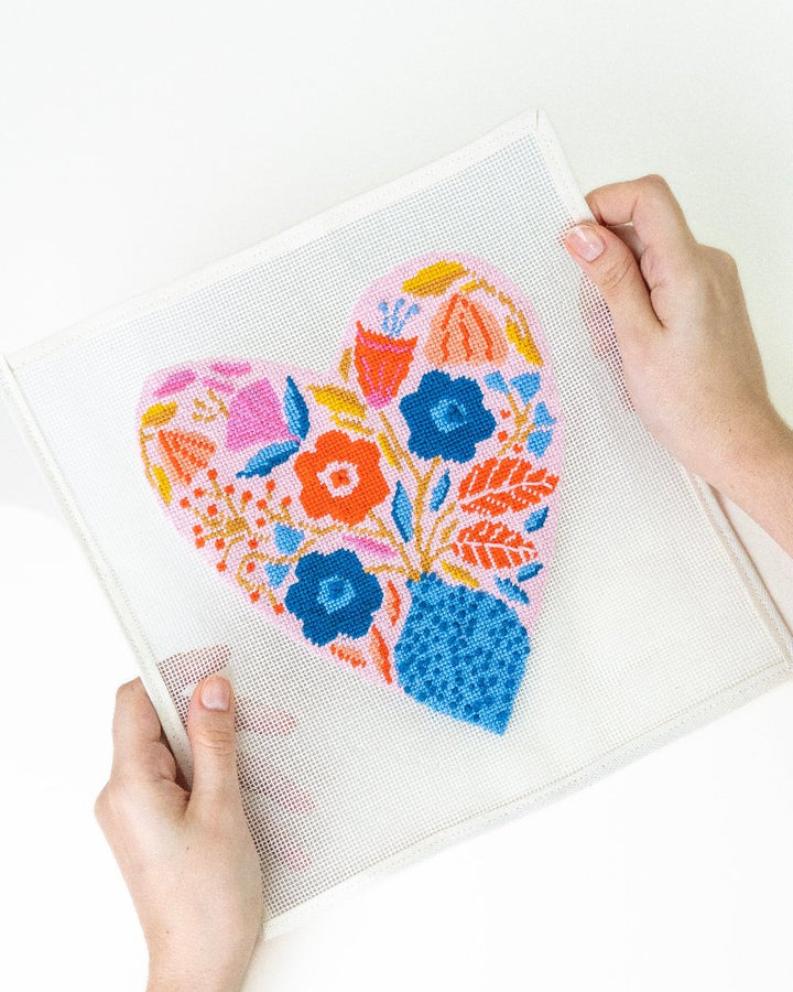 A needlepoint kit depicting a heart-shaped canvas with a floral design being held by a pair of hands.