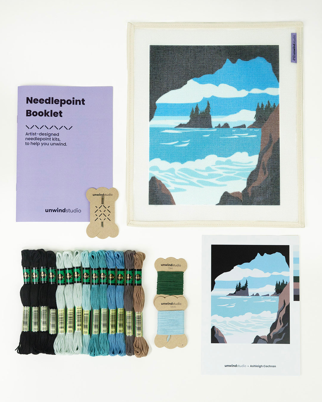 Needlepoint kit with a mountain and ocean design on a light blue background. Black, three shades of blues, grey, brown and green threads. A postcard with the design and a needlepoint booklet with two embroidery needles next to it.