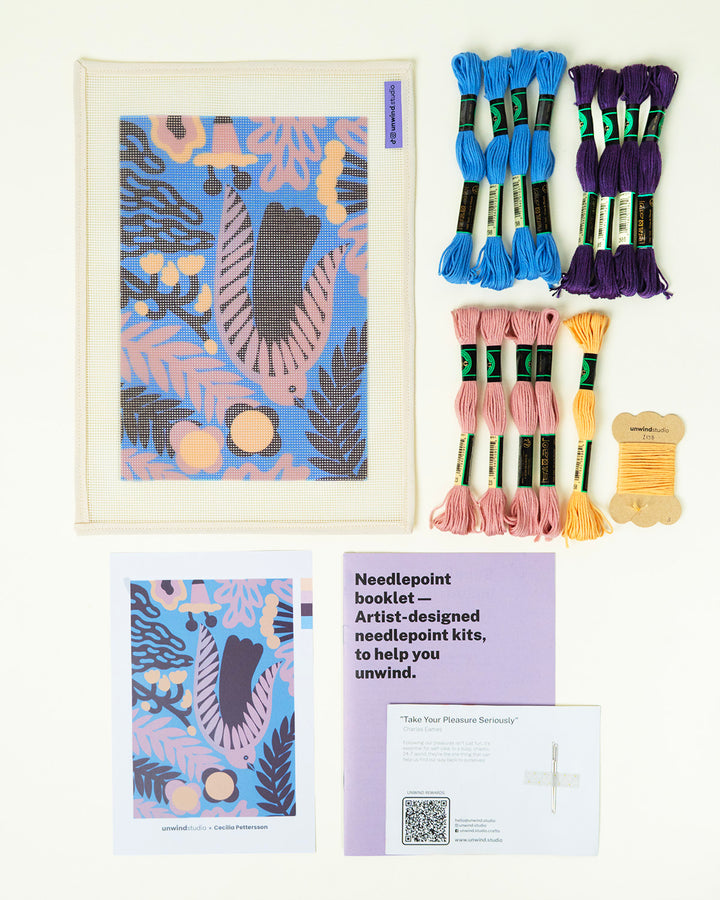 Needlepoint kit with thread skeins, a framed artwork, and a booklet on a white background