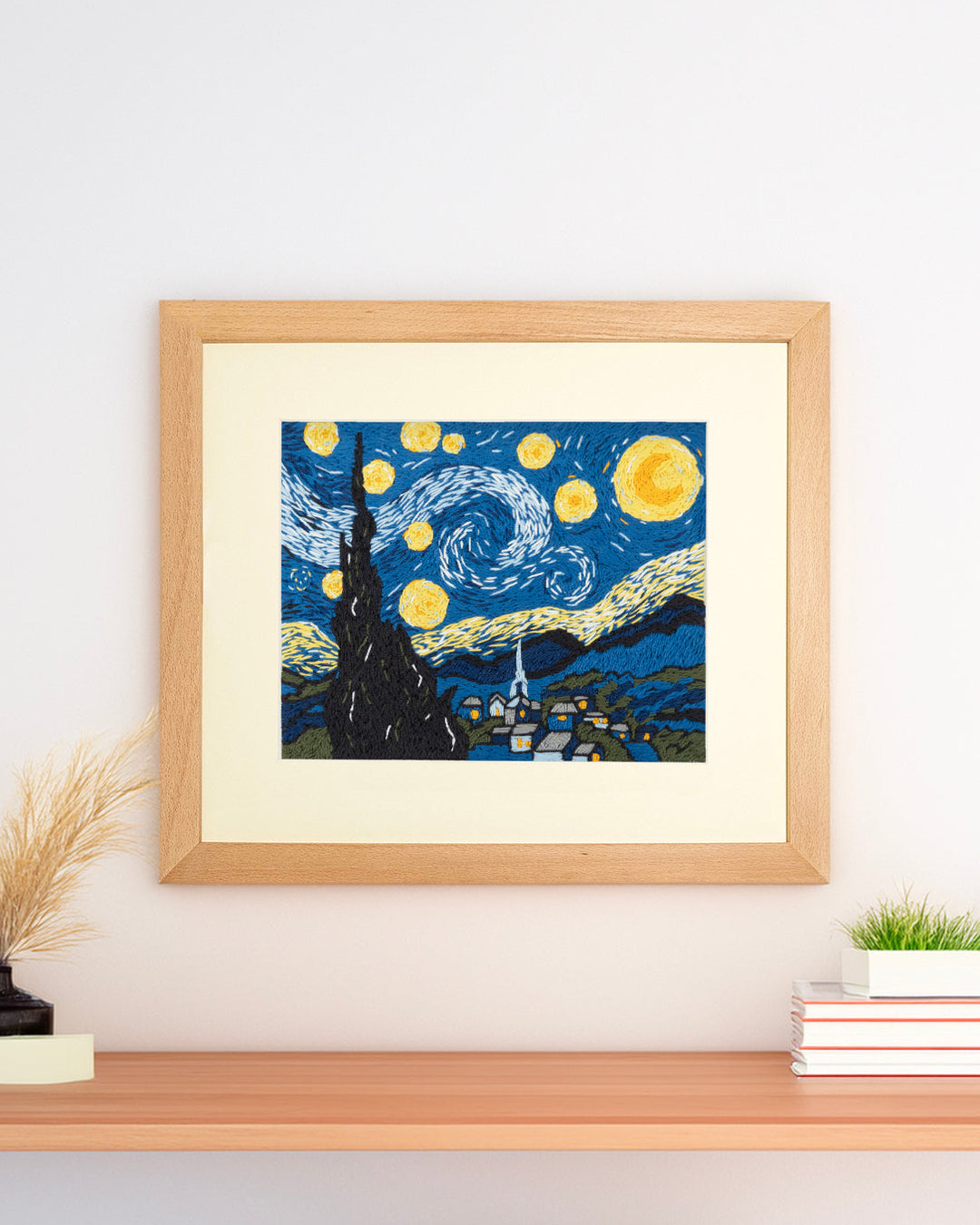 Framed needlepoint artwork of 'Starry Night' by Vincent van Gogh on a wall above a shelf.