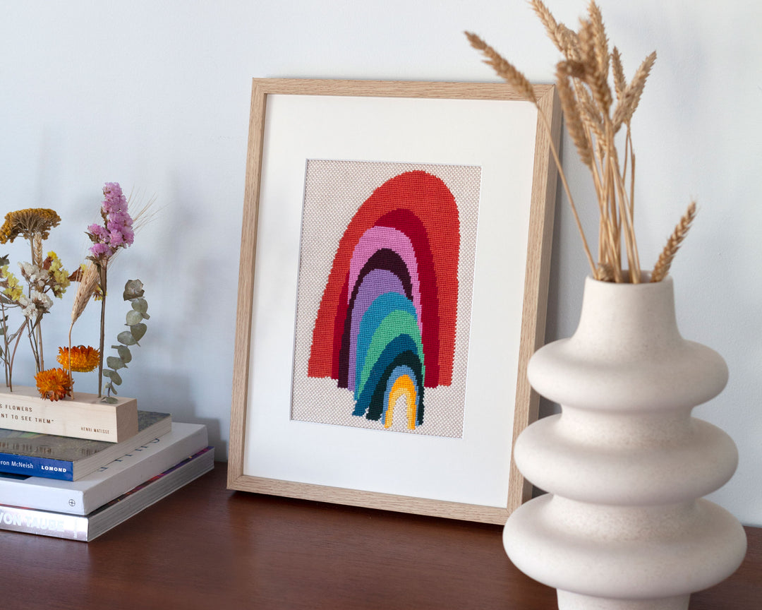 Framed needlepoint rainbow illustration next to vase and books