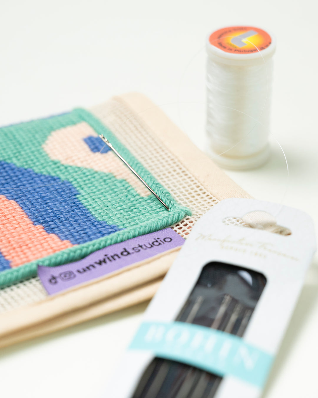 a diagonal view of the Bohin France set of Sewing Needles and a single needle placed on top of a needlepoint canvas