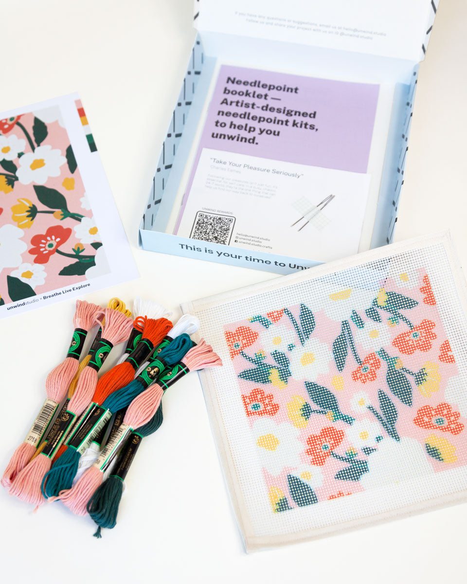needlepoint kit with small canvas with illustration of floral pattern with flowers and leaves, next to threads and other accessories and a postcard with the original illustration by Unwind Studio