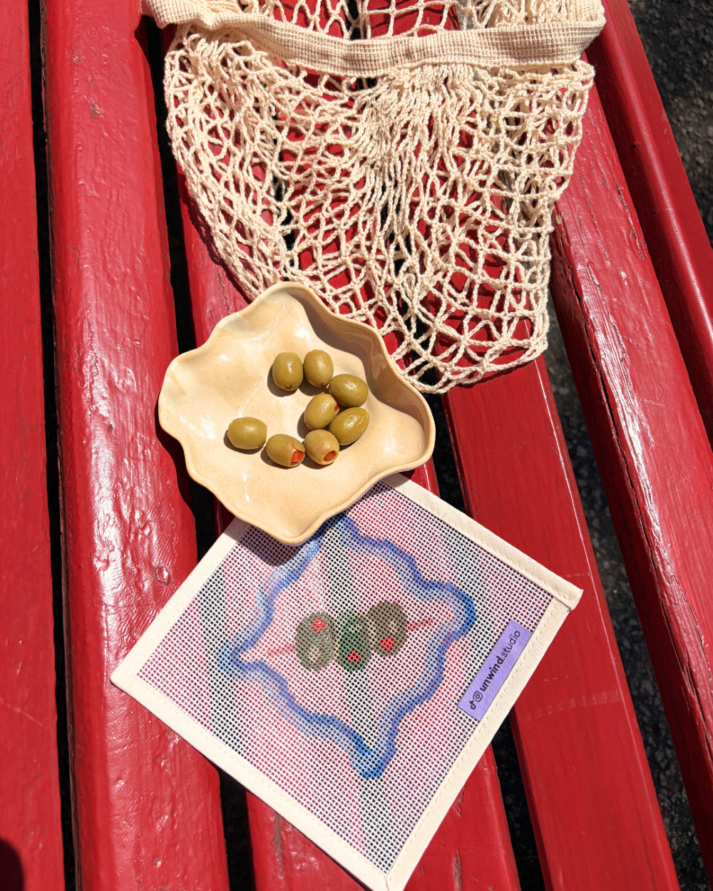 Close-up shot of the Unwind Studio coaster from the italian summer collection with an olive on a toothpick placed on a red wood park bench.