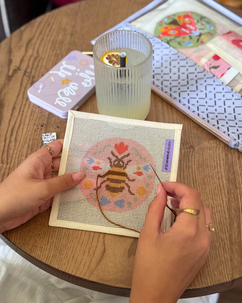 A person's hands are shown needlepointing a bee design on a canvas over a wooden table. A glass of iced drink and other crafting items like a project bag are visible in the background.