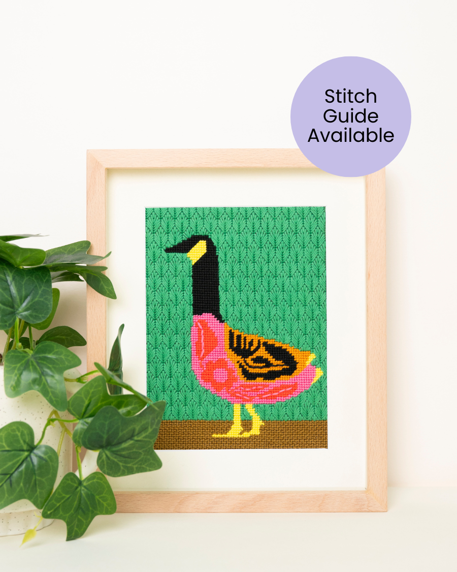 Folk Goose Needlepoint kit by unwind studio placed in a wood frame with a decorative plant.