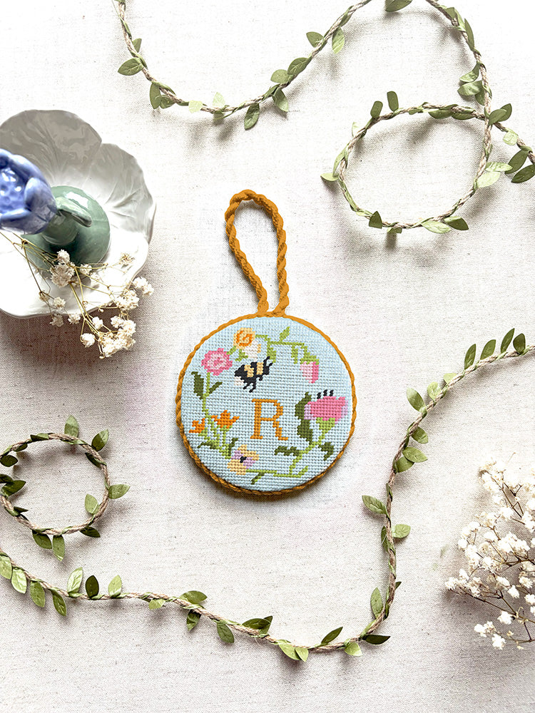 Top-down view of a finished round needlepoint ornament with a braided hanging loop, showcasing Unwind Studio's Personalizable Floral Trio design. The Garden Bee canvas features a colorful spring floral wreath with a tiny insect nestled in the petals and a personalized monogram letter hand-stitched right in the center. The ornament is styled on a neutral linen backdrop, surrounded by delicate faux green leaf vines, dried white baby's breath flowers, and a pastel purple ceramic candlestick holder. 