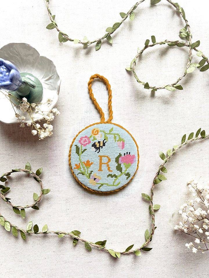 Top-down view of a finished round needlepoint ornament with a braided hanging loop, showcasing Unwind Studio's Personalizable Floral Trio design. The Garden Bee canvas features a colorful spring floral wreath with a tiny insect nestled in the petals and a personalized monogram letter hand-stitched right in the center. The ornament is styled on a neutral linen backdrop, surrounded by delicate faux green leaf vines, dried white baby's breath flowers, and a pastel purple ceramic candlestick holder. 