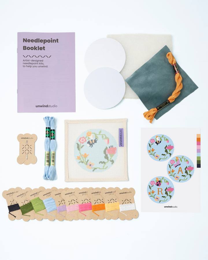 Flat view of the Garden Bee Needlepoint Ornament by Unwind Studio with everything that comes with the kit: an instructional booklet, needle, dmc soft cotton threads, finishing materials and a graphic flyer