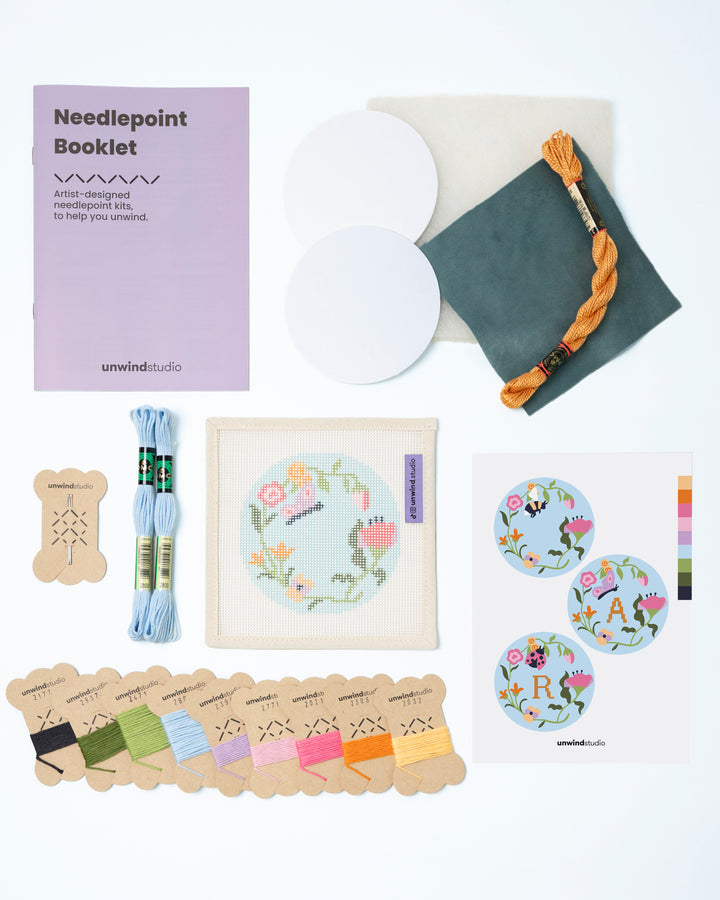 Flat view of the Garden Butterfly Needlepoint Ornament by Unwind Studio with everything that comes with the kit: an instructional booklet, needle, dmc soft cotton threads, finishing materials and a graphic flyer