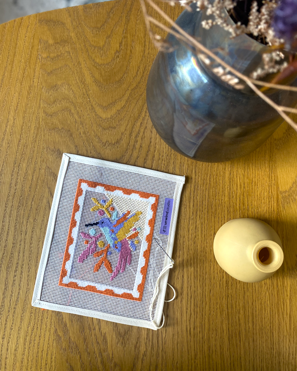 Hummingbird needlepoint canvas kit with colorful bird design and orange border, stitched on white mesh canvas, viewed from above.