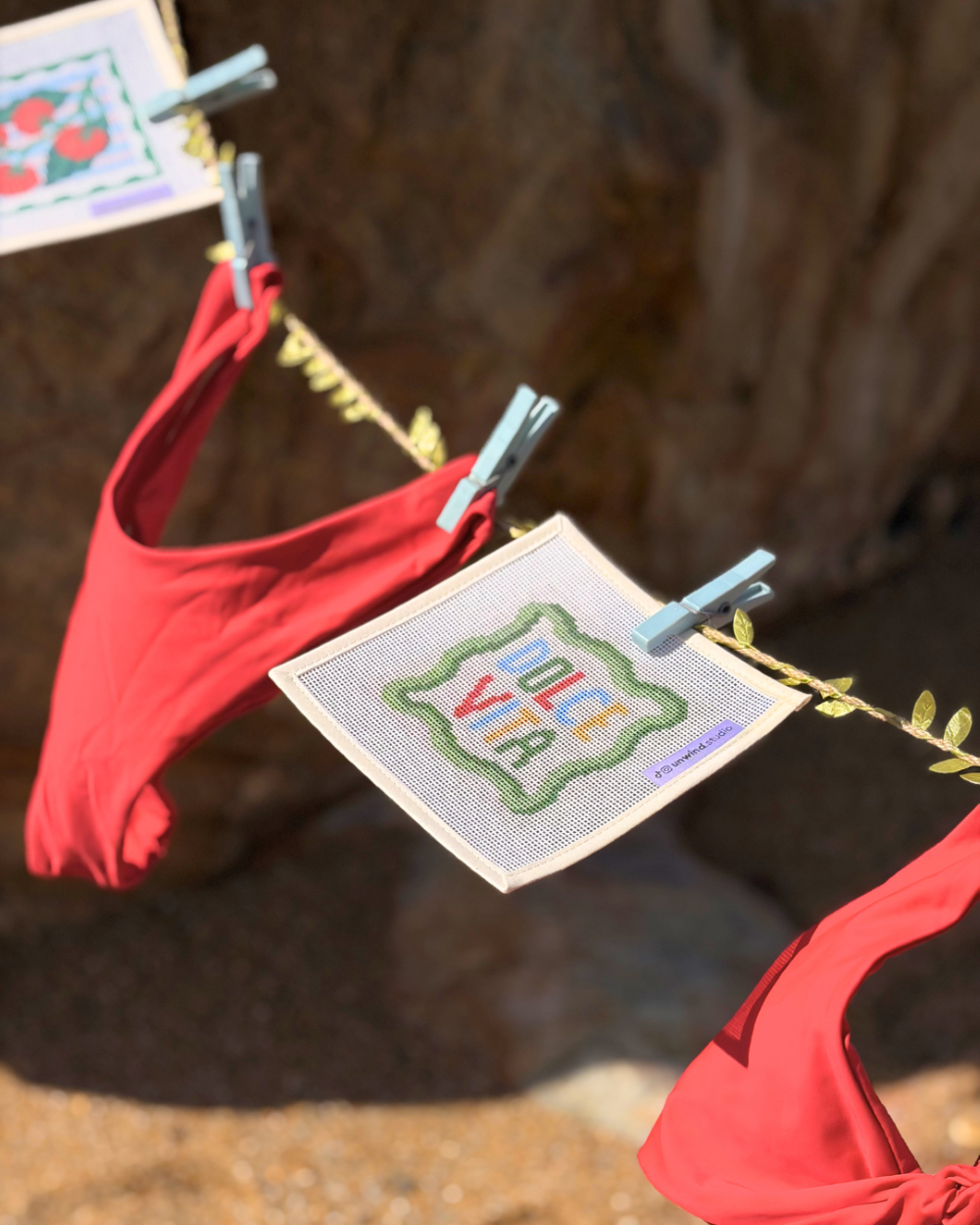 A close-up shot of an Unwind Studio needlepoint canvas featuring the "Dolce Vita" typographic design with a green scalloped border, hanging from a clothesline with a light blue clothespin.