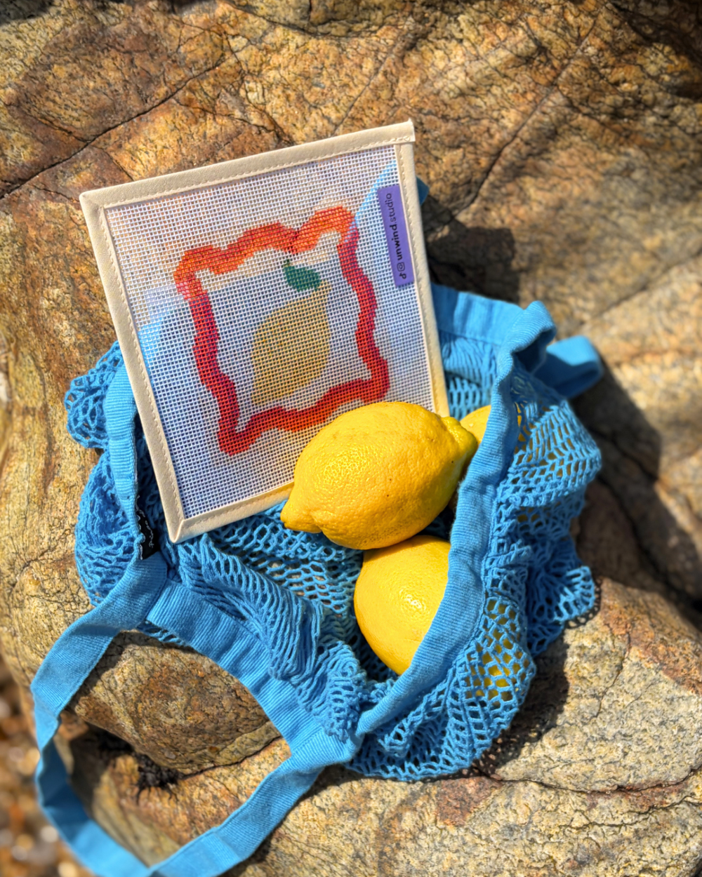 A close-up shot of an Unwind Studio needlepoint canvas featuring the Lemon design with a red scalloped border in a blue fruit bag placed on a beach rock.