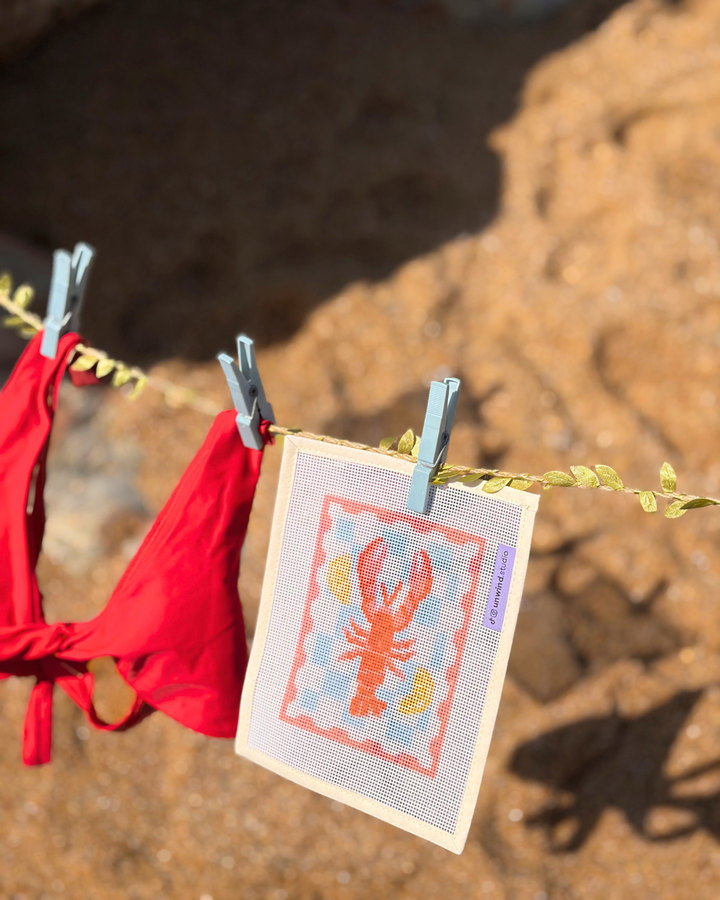 close-up shot of an Unwind Studio needlepoint canvas featuring the Lobster Stamp design hanging from a clothesline with a light blue clothespin on a beach.