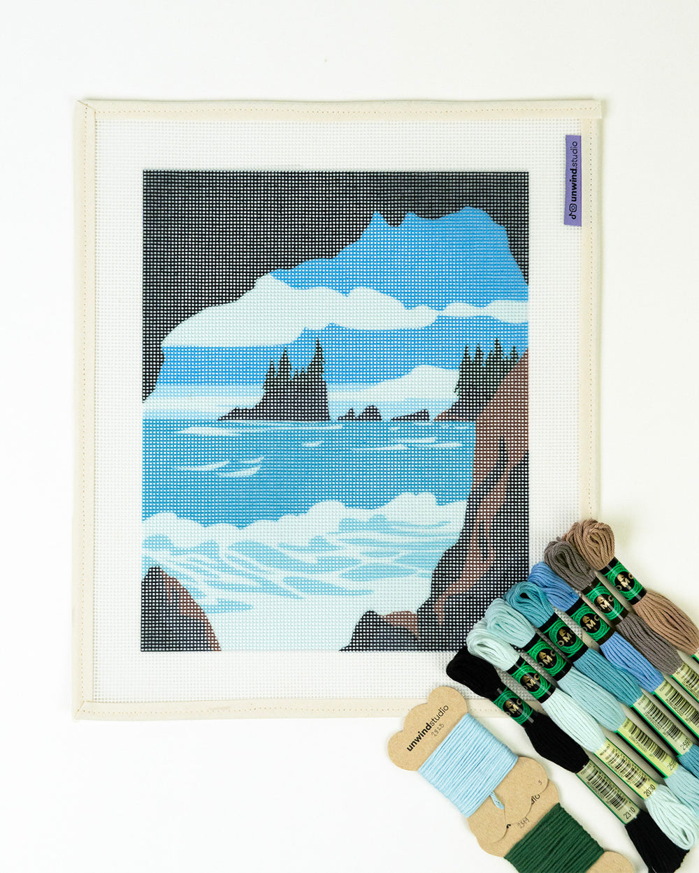 Needlepoint kit with a mountain and ocean design on a light blue background, with blue, black, green and brown threads on top.
