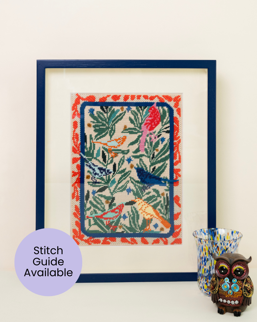 the birds needlepoint kit by unwind studio framed in a blue wood frame with a painted glass and a owl as decorative elements.