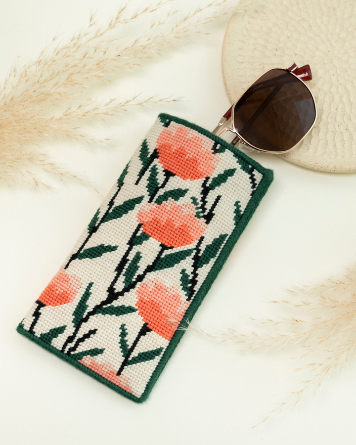 up-close shot of unwind studio about me sunglasses needlepoint case in a white background with dry flowers and the glasses coming out.
