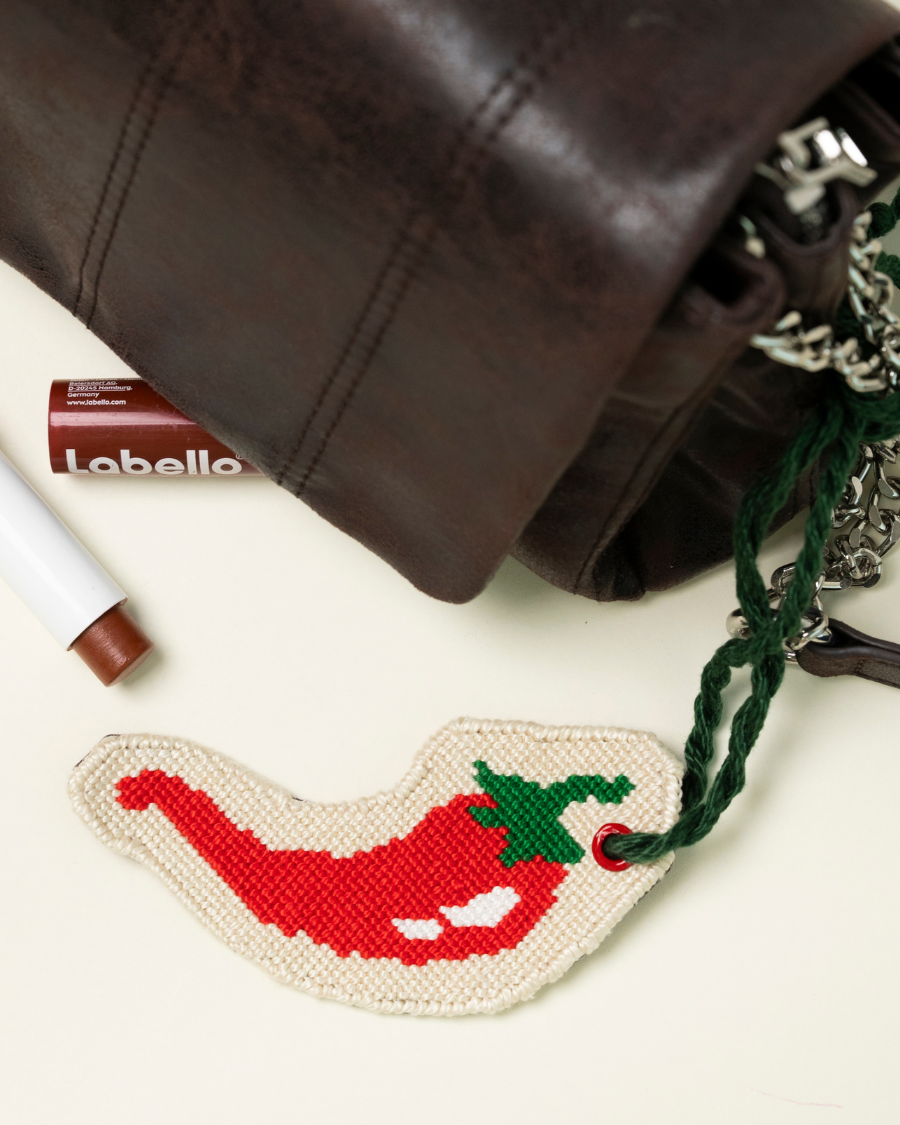 chili pepper needlepoint bag charm by unwind studio placed on a brown bag with a lipstick by its side
