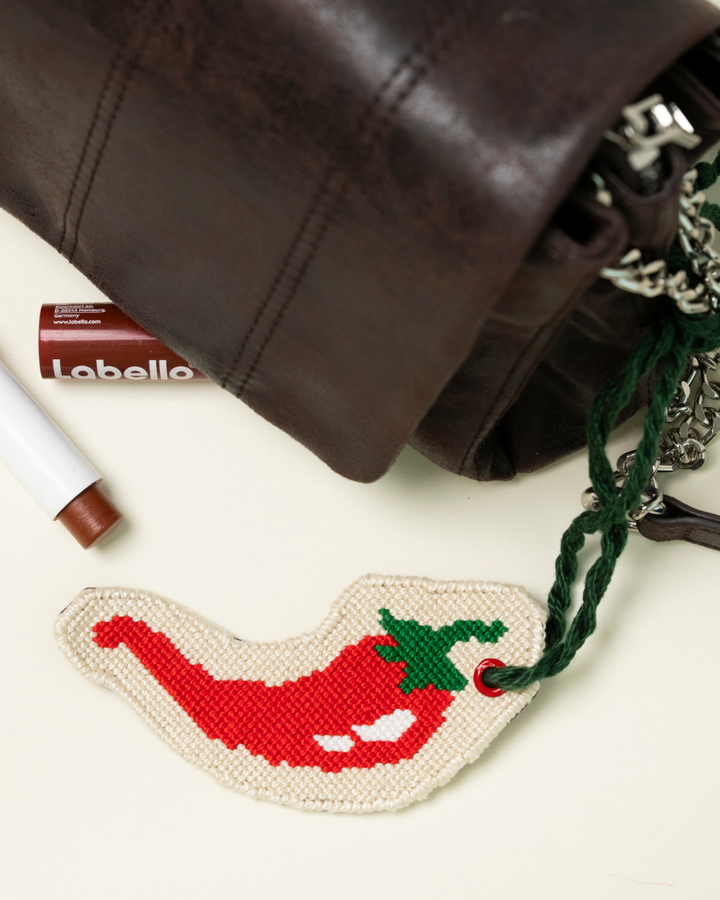 chili pepper needlepoint bag charm by unwind studio placed on a brown bag with a lipstick by its side