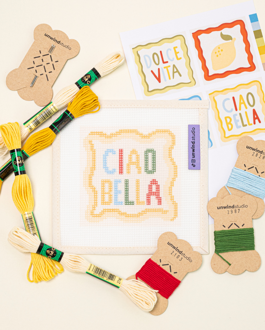 flat lay of the ciao bella needlepoint coaster paired with the matching DMC Soft Cotton Threads, needle and graphic flyer.