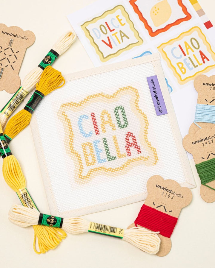 diagonal flat lay of the ciao bella needlepoint coaster paired with the matching DMC Soft Cotton Threads and graphic flyer.