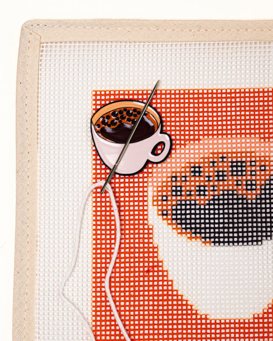 close shot of unwind studio needle minder coffee (bica) magnet placed on a matching coffee (bica) coaster needlepoint canvas with a needle with a white thread on top.

