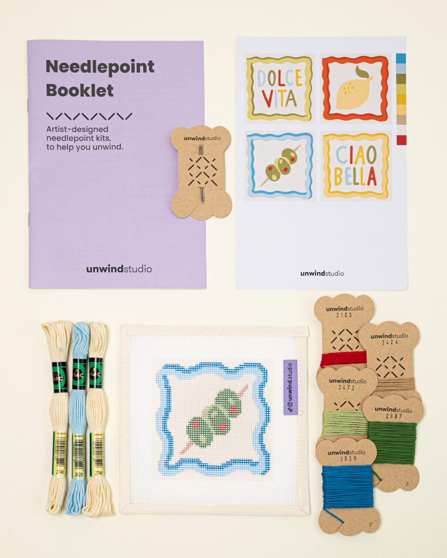 Top-down view of the dirty martini Coaster needlepoint kit including canvases, DMC Soft Cotton thread, flyer, and an instructional booklet.


