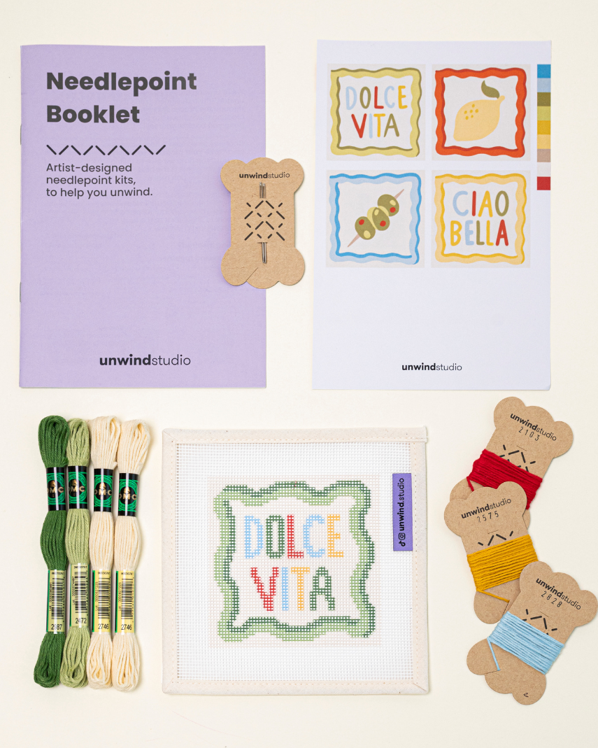 Top-down view of the dolce vita Coaster  needlepoint kit including canvases, DMC Soft Cotton thread, flyer, and an instructional booklet.

