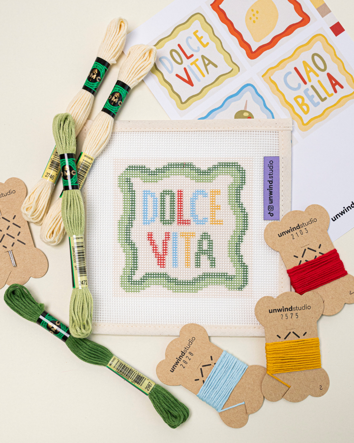 flat lay of the unwind studio dolce vita needlepoint coaster paired with matching needlepoint threads and graphic flyer.