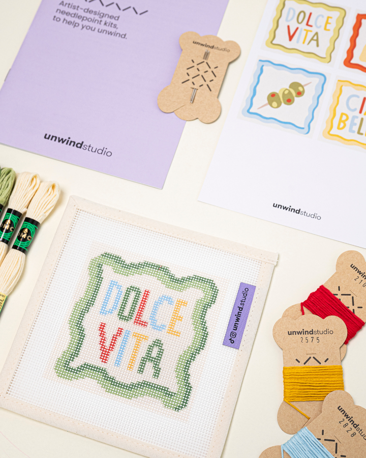 diagonal view of the dolce vita Coaster set needlepoint kit including canvases, DMC Soft Cotton thread, flyer, and a instructional booklet.