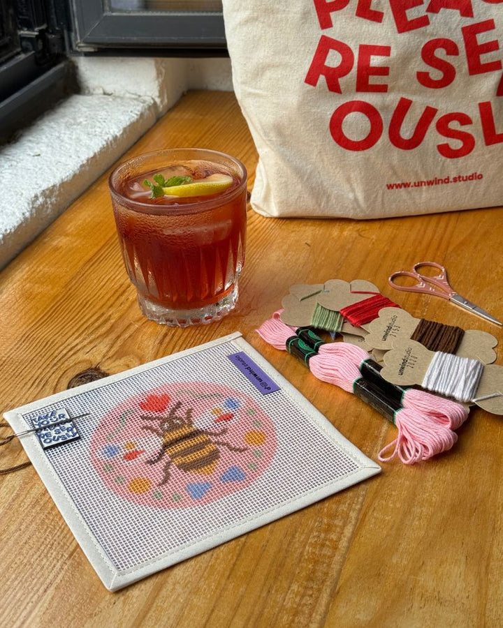 A cozy craft setup on a wooden table featuring a completed bee needlepoint design in a square frame, alongside spools of embroidery floss, scissors, wooden beads, and a branded canvas tote bag from Unwind Studio.
