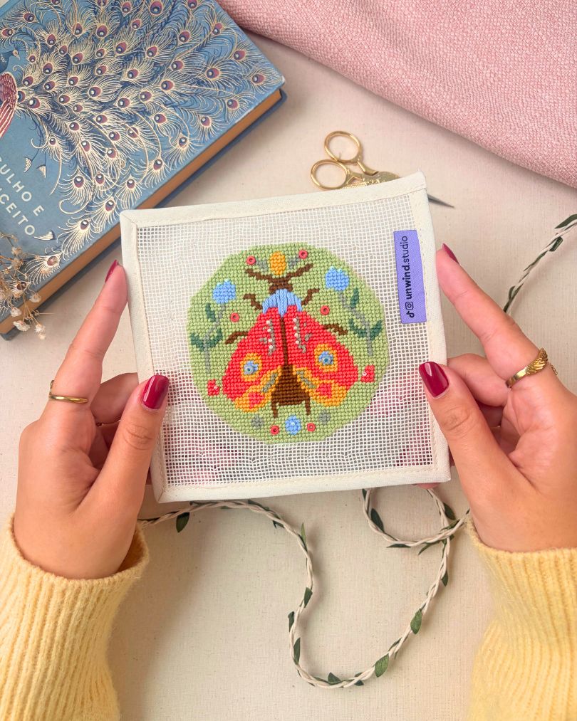 A person holds an ornament needlepoint canvas featuring a colorful moth design in the center. The hands have dark red nail polish and gold rings, and the background includes a blue peacock-patterned book, a pink fabric, gold scissors, and a green-and-white bunting ribbon.