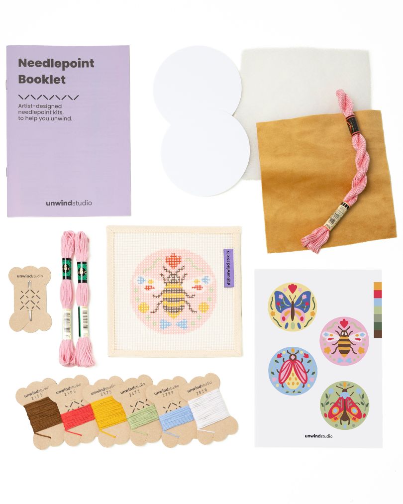 Flat view of the Folk Bee Needlepoint Ornament by Unwind Studio with everything that comes with the kit: an instructional booklet, needle, dmc soft cotton threads, finishing materials and a graphic flyer
