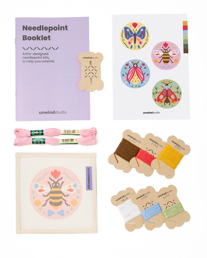 Flat view of the Folk Bee Needlepoint Ornament by Unwind Studio with an instructional booklet, needle, dmc soft cotton threads and a graphic flyer