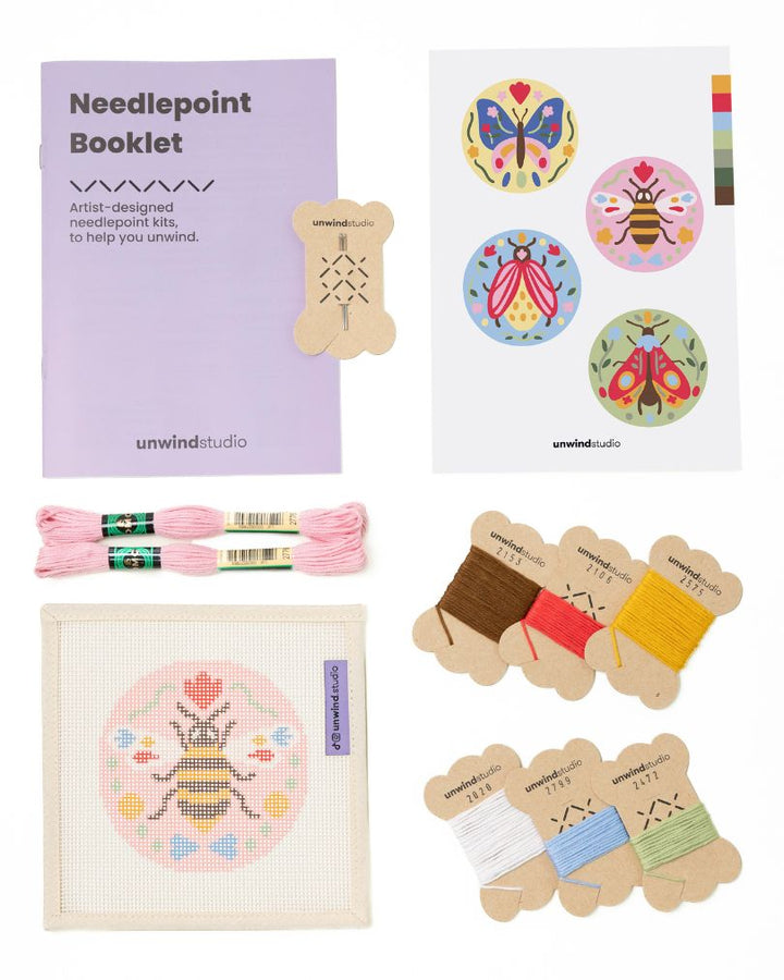 Flat view of the Folk Bee Needlepoint Ornament by Unwind Studio with an instructional booklet, needle, dmc soft cotton threads and a graphic flyer
