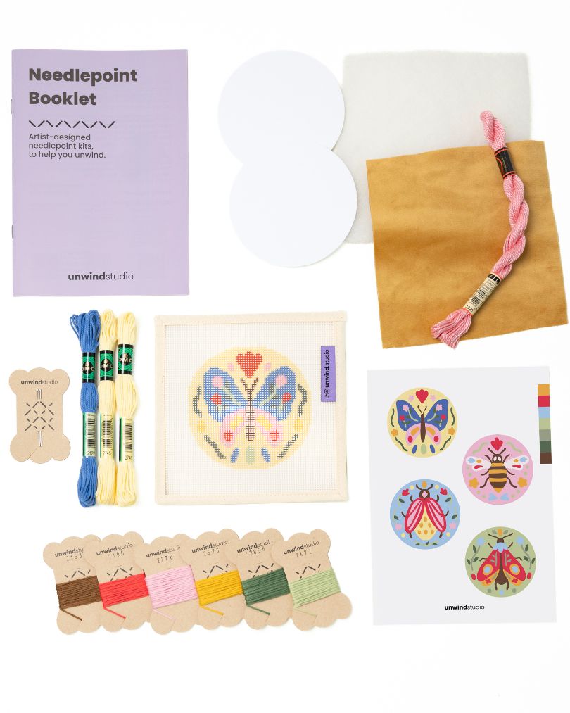Flat view of the Folk butterfly Needlepoint Ornament by Unwind Studio with everything that comes with the kit: an instructional booklet, needle, dmc soft cotton threads, finishing materials and a graphic flyer
