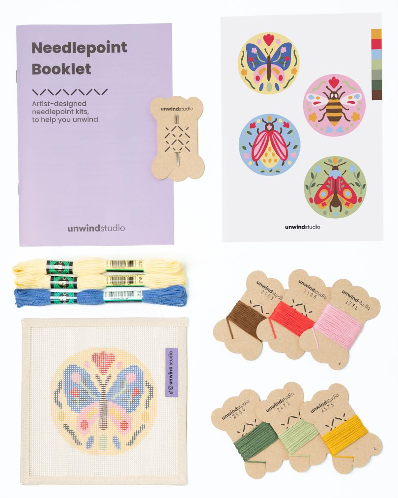 Flat view of the Folk butterfly Needlepoint Ornament by Unwind Studio with an instructional booklet, needle, dmc soft cotton threads and a graphic flyer
