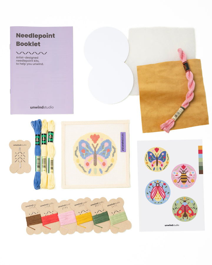 Flat view of the Folk Butterfly Needlepoint Ornament by Unwind Studio with everything that comes with the kit: an instructional booklet, needle, dmc soft cotton threads, finishing materials and a graphic flyer

