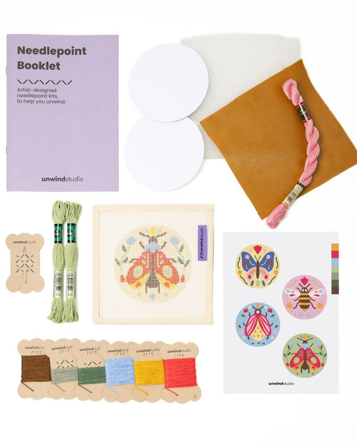 Flat view of the Folk moth Needlepoint Ornament by Unwind Studio with everything that comes with the kit: an instructional booklet, needle, dmc soft cotton threads, finishing materials and a graphic flyer
