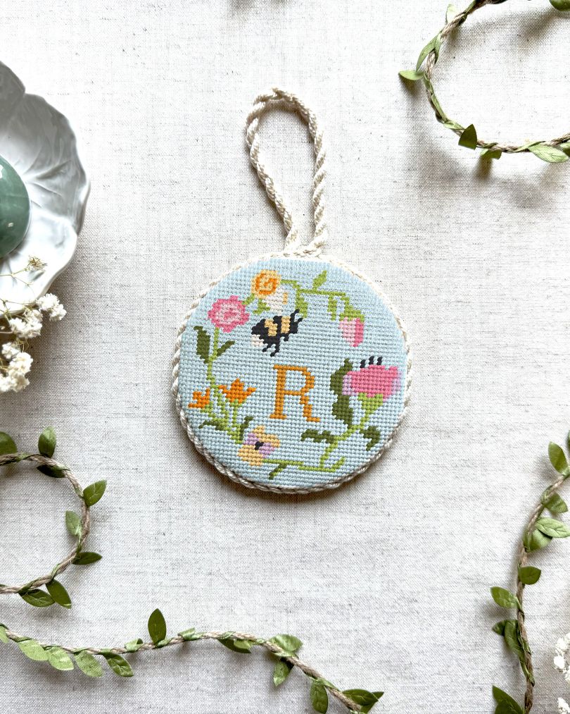 Top-down view of a finished round needlepoint ornament with a braided hanging loop, showcasing Unwind Studio's Personalizable Floral Trio design. The Garden Bee canvas features a colorful spring floral wreath with a tiny insect nestled in the petals and a personalized monogram letter hand-stitched right in the center. The ornament is styled on a neutral linen backdrop, surrounded by delicate faux green leaf vines, dried white baby's breath flowers, and a pastel purple ceramic candlestick holder.
