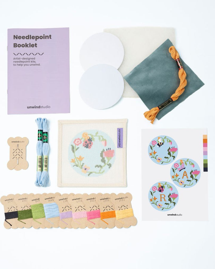 Flat view of the Garden ladybug Needlepoint Ornament by Unwind Studio with everything that comes with the kit: an instructional booklet, needle, dmc soft cotton threads, finishing materials and a graphic flyer

