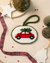 An oval needlepoint ornament featuring a red car with a green Christmas tree on its roof, surrounded by holiday-themed props like a plaid ribbon, star and tree tags, and seashells on a beige fabric background.