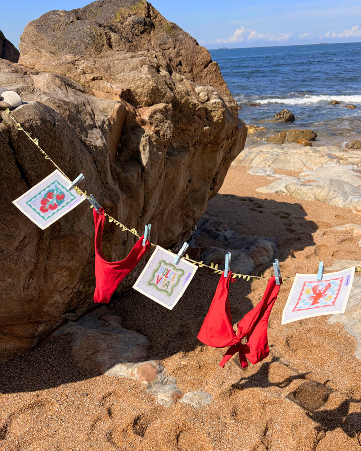 A lifestyle shot of an Unwind Studio needlepoint canvases featuring the Italian Summer Collection hanging from a clothesline with a light blue clothespin and a red bikini on a beach.