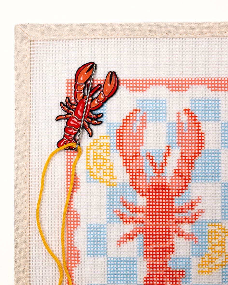 close shot of unwind studio needle minder lobster magnet placed on a matching lobster stamp needlepoint canvas with a needle with a yellow thread on top.