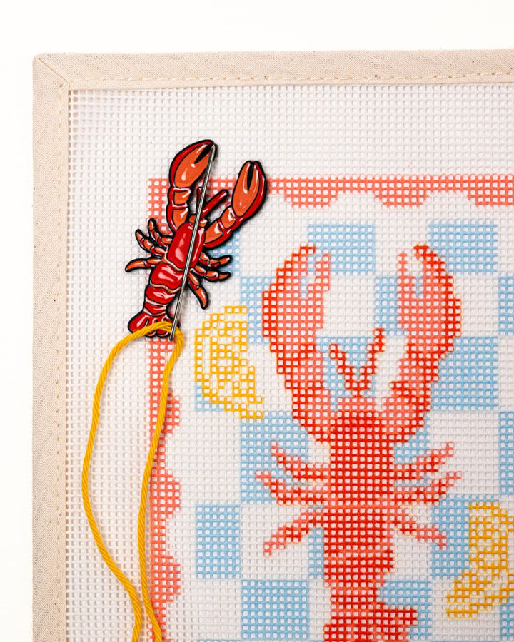 close shot of unwind studio needle minder lobster magnet placed on a matching lobster stamp needlepoint canvas with a needle with a yellow thread on top.