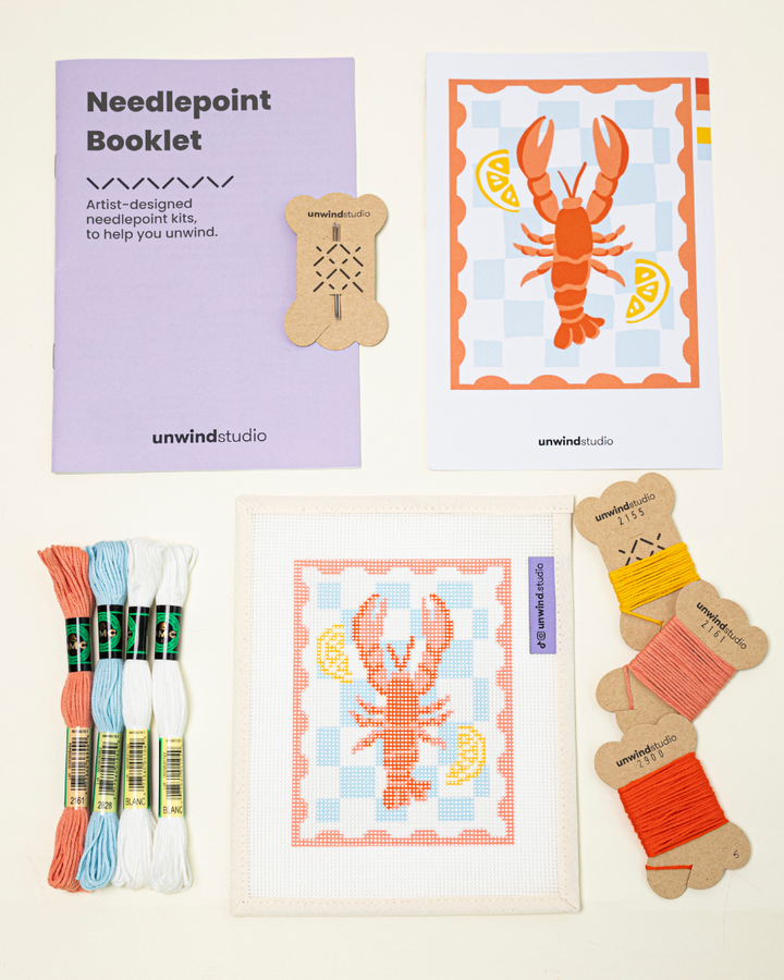 Top-down view of the lobster stamp needlepoint kit including canvases, DMC Soft Cotton thread, flyer, and an instructional booklet.