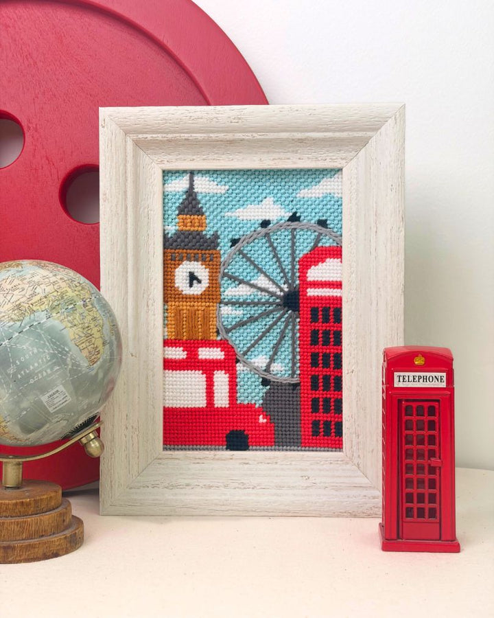 A framed cross-stitch artwork depicting London landmarks, including Big Ben, a red double-decker bus, a red telephone booth, and a Ferris wheel, styled on a tabletop with a globe and a red decorative object in the background.