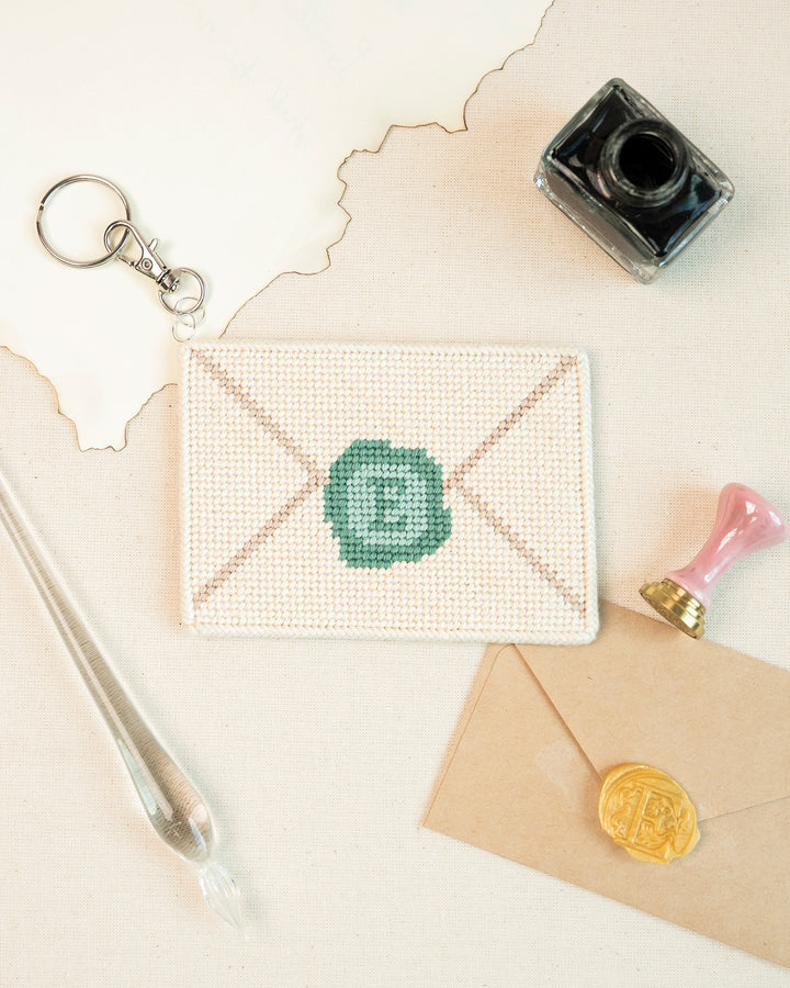 Love Letter needlepoint Bag Charm finished paired next to a look-alike envelop, a quill pen and paint.