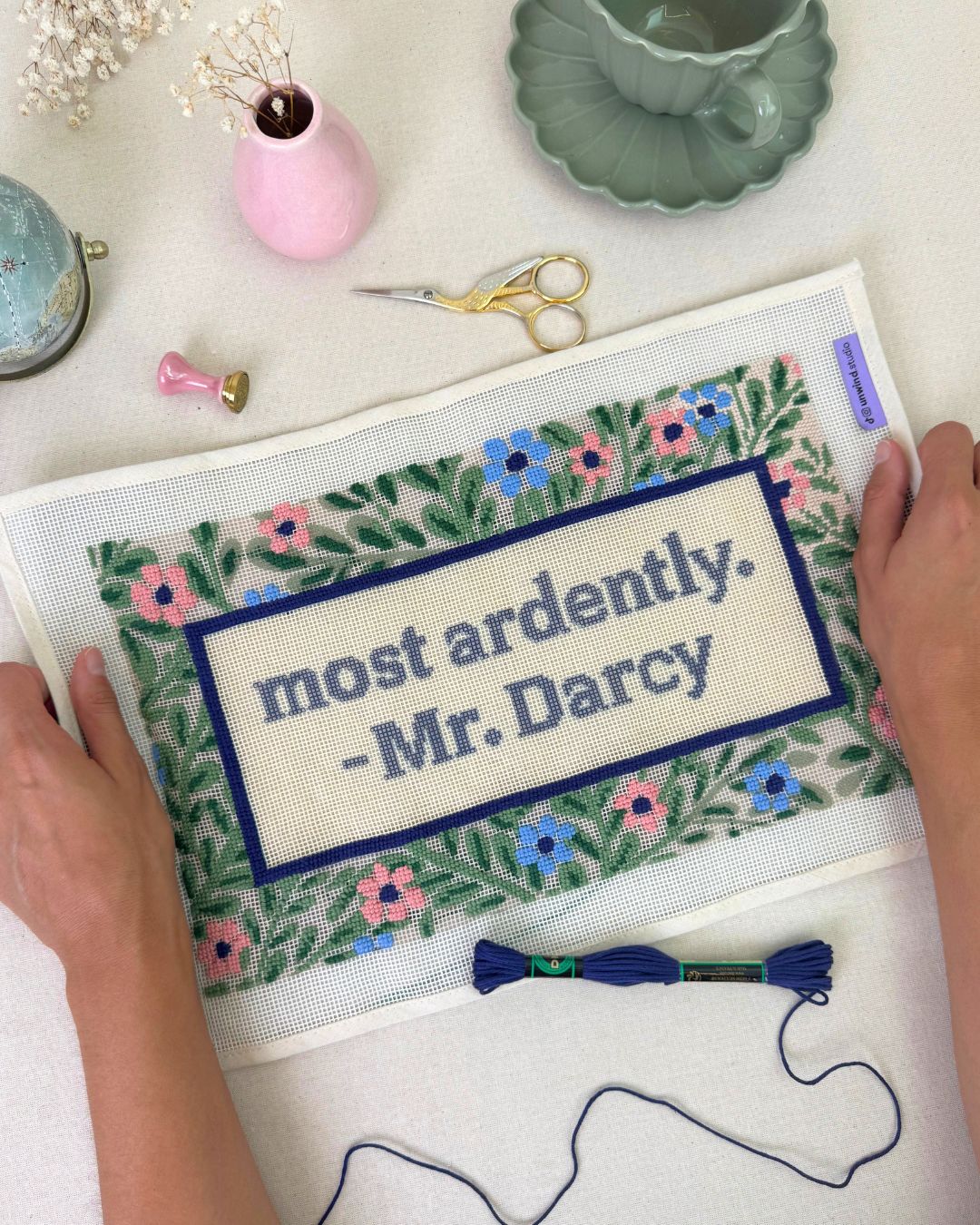 Most Ardently Needlepoint cushion canvas in progress with matching regency decor elements such as a tea cup, a letter stamp, a little globe and threads